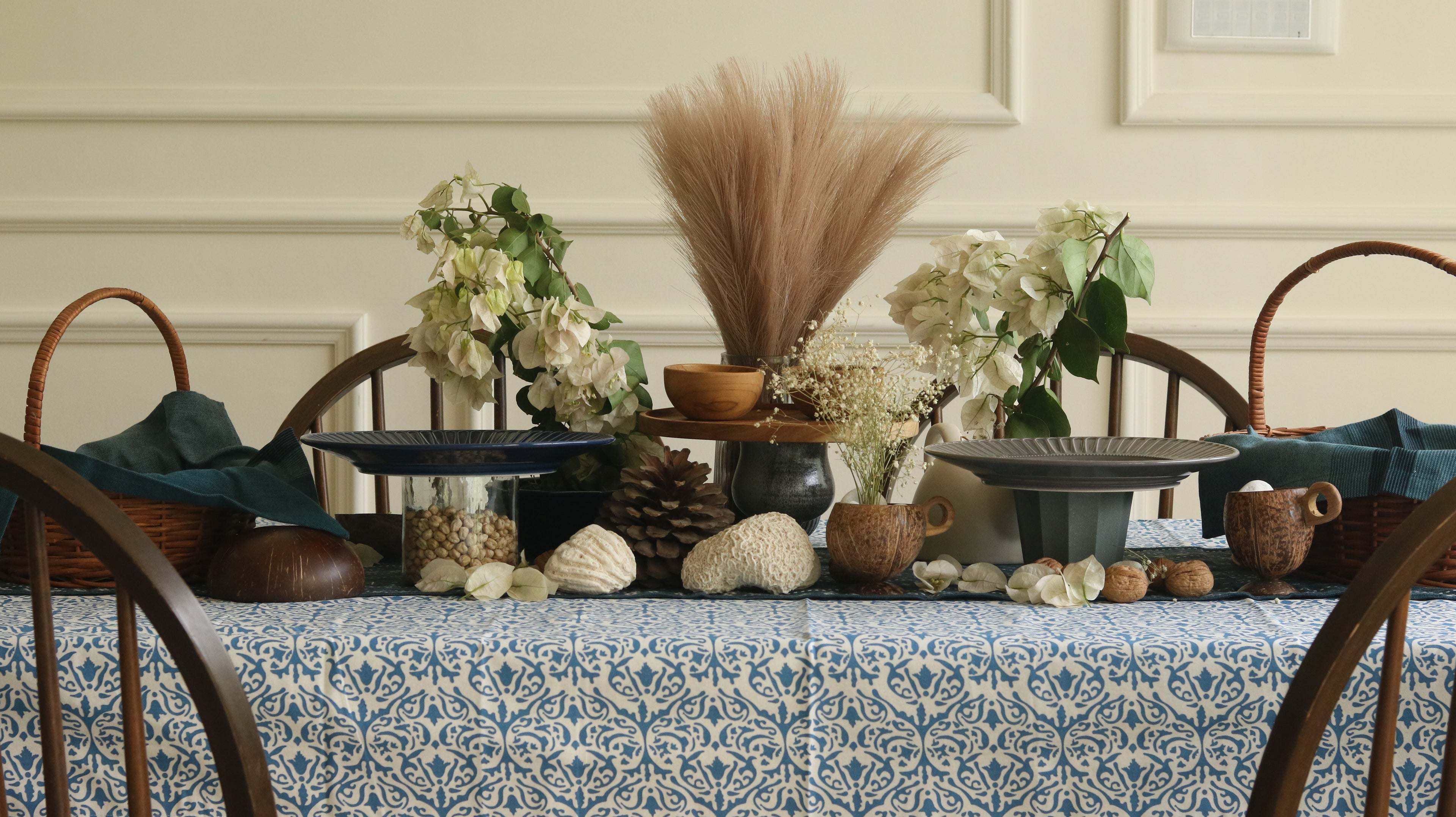 A blue and white patterned tablecloth styled with wooden chairs and serving stands with rustic center piece features white bougainvillea, dried pampas grass, and coconut shell cups.