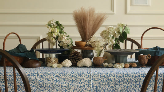 A blue and white patterned tablecloth styled with wooden chairs and serving stands with rustic center piece features white bougainvillea, dried pampas grass, and coconut shell cups.