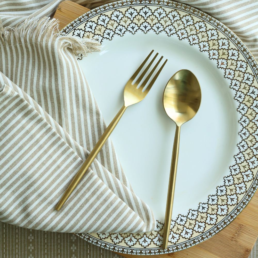 Matte gold dinner spoon and fork laid across a plate and striped napkin