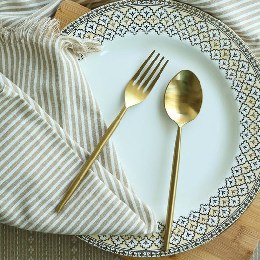 Matte gold dinner spoon and fork laid across a plate and striped napkin
