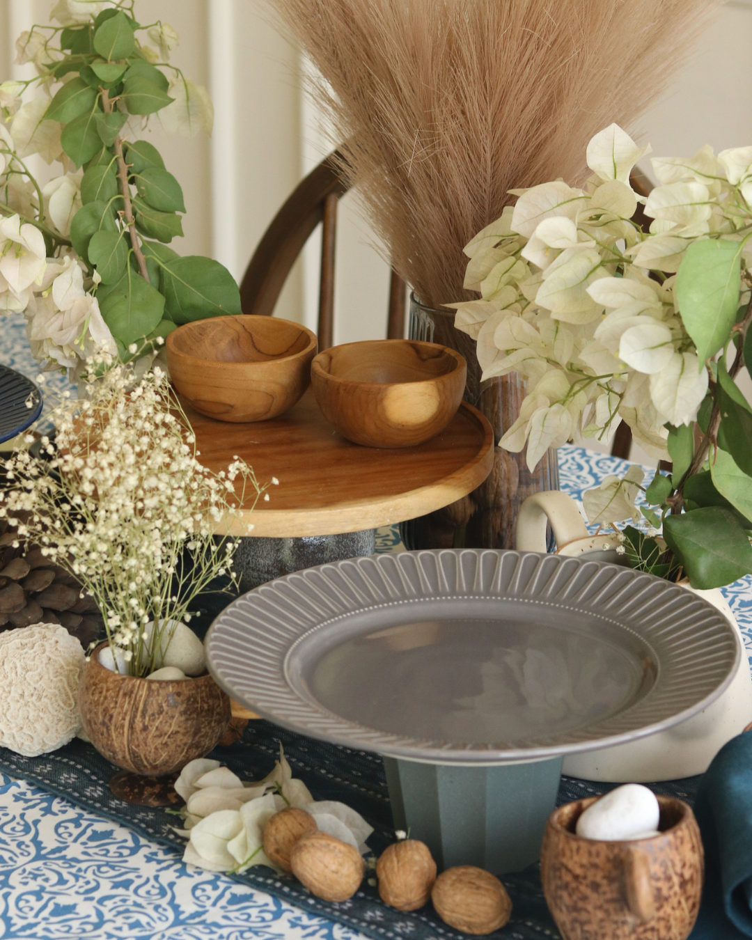A styled tabletop arrangement featuring a large ribbed gray pedestal plate in the foreground, wooden bowls on a round wooden tray, dried flowers, white blossoms, and blue-and-white patterned textiles in a warm home setting.