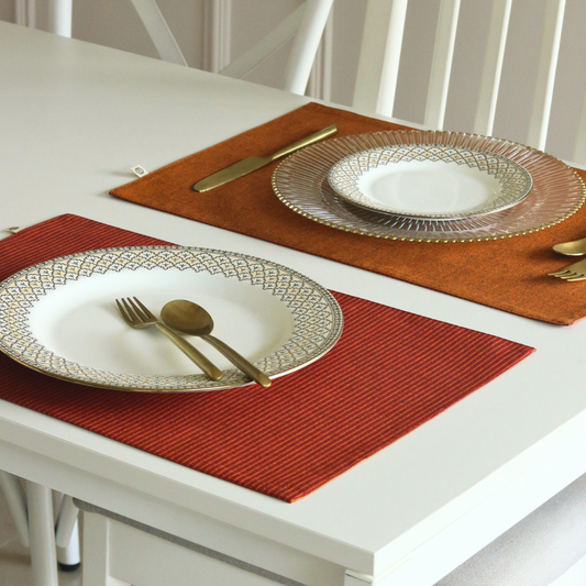 Two place settings on a white table, each with a white patterned plate, a clear scalloped charger plate, gold-toned cutlery, and a rectangular placemat in rust red or burnt orange. No people or animals are present.