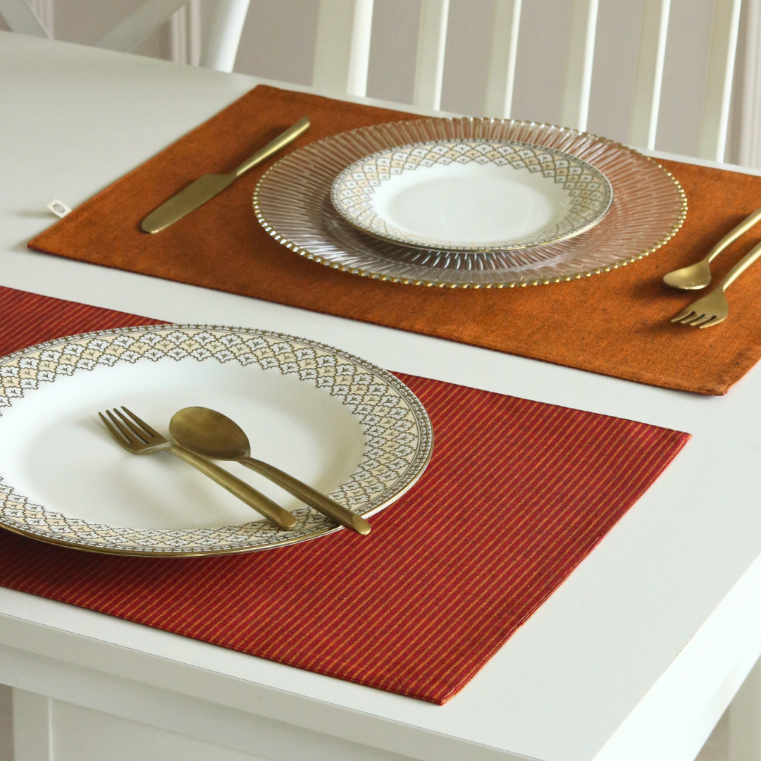 Two woven placemats on a white table, one rust orange and one red, each styled with a white patterned plate, a clear textured charger plate, and gold-toned cutlery.