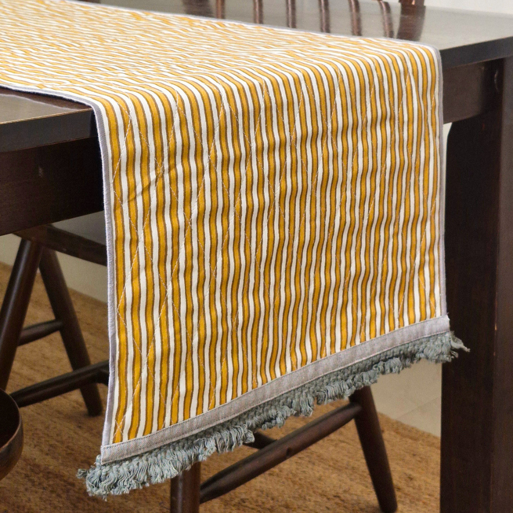 An ochre and white 6 seater reversible runner hanging over a wooden dining table with grey tassels.