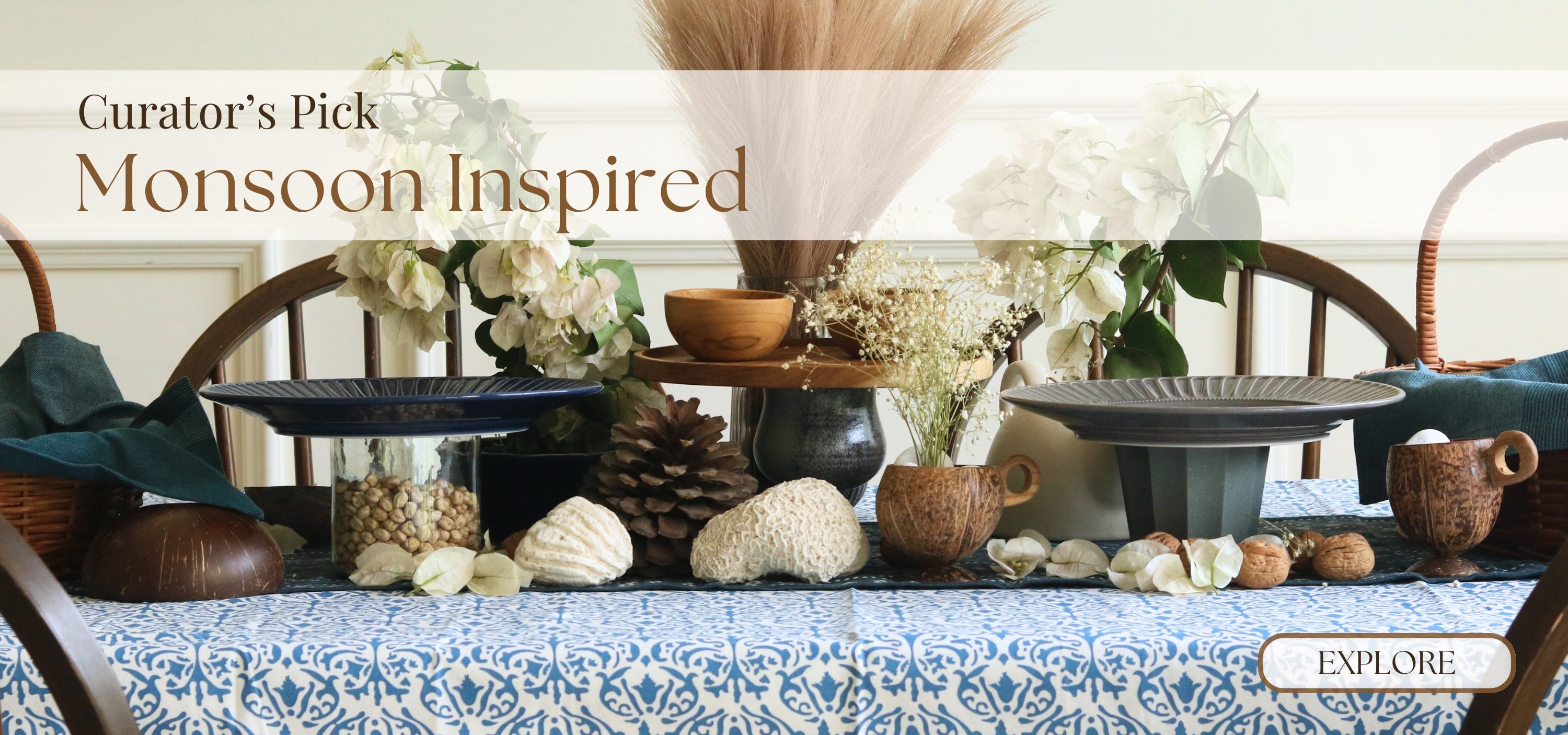 A styled tabletop display with monsoon-inspired decor, including a blue patterned tablecloth, woven baskets, ceramic vessels, shells, dried grasses, and floral arrangements. Large text at the top reads 'Curator's Pick Monsoon Inspired,' and a small 'EXPLORE' button appears in the lower right.